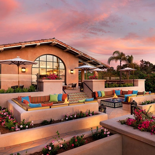Outdoor lounge area at Rancho Valencia Resort during sunset, featuring colorful cushions, fire pits, flowering garden beds, and Mediterranean-style architecture