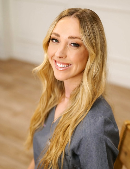 Sam Bausback, R.N., B.S.N., surgical nurse, smiling and seated indoors, wearing a gray scrub top with long blonde hair styled in loose waves