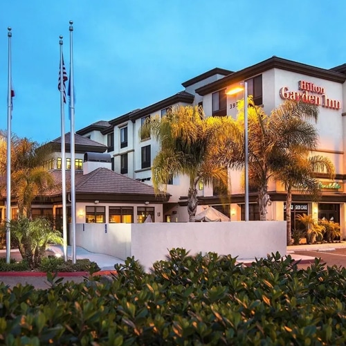 Exterior of Hilton Garden Inn San Diego Del Mar in Carmel Valley, featuring palm trees and the hotel entrance at dusk.